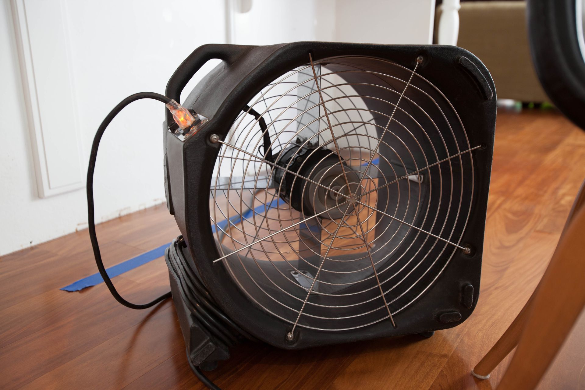 A black air mover fan sits on a wooden floor, with a blue painter's tape line nearby.