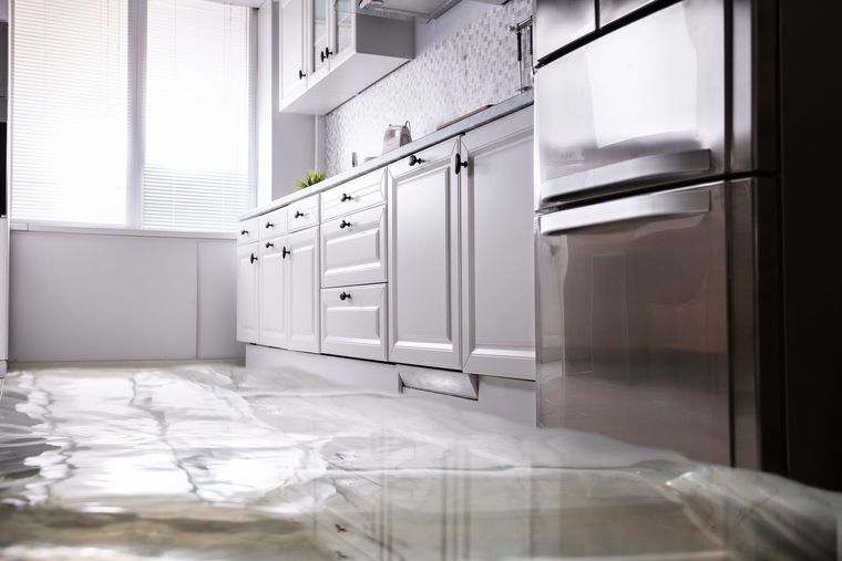 A flooded kitchen floor covered in shallow standing water in front of white cabinets and a stainless steel refrigerator.