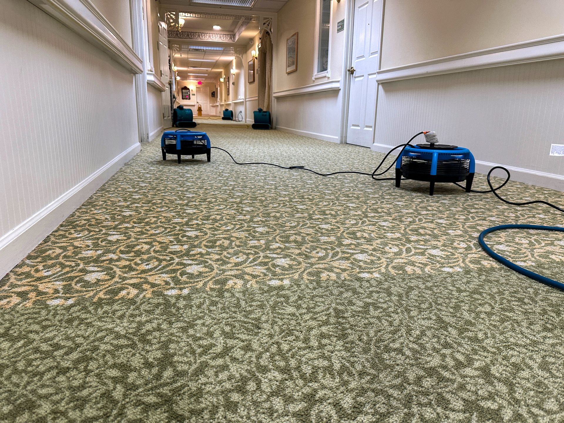 A hotel hallway with industrial drying fans placed on a patterned green carpet, likely indicating water damage repair.