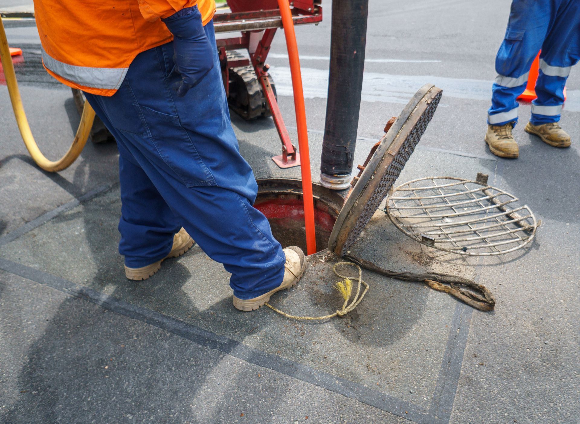 Utility workers stand over an open manhole on an asphalt road, using a hose and equipment for underground work.