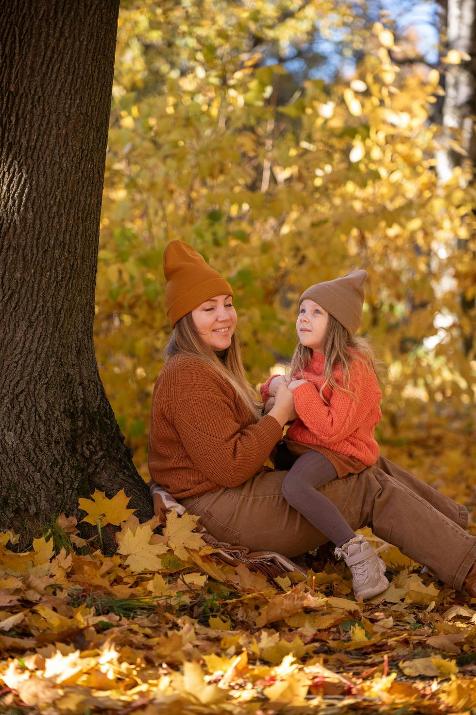Mutter und Kind in passenden orangefarbenen Pullovern und Mützen sitzen neben einem Baum im Herbstlaub.