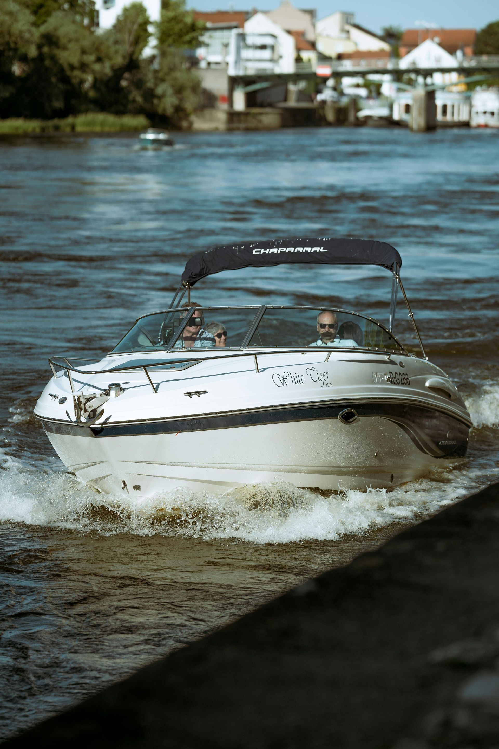 Motorboat on a river, two people visible. White boat with black canopy. Buildings in the background.