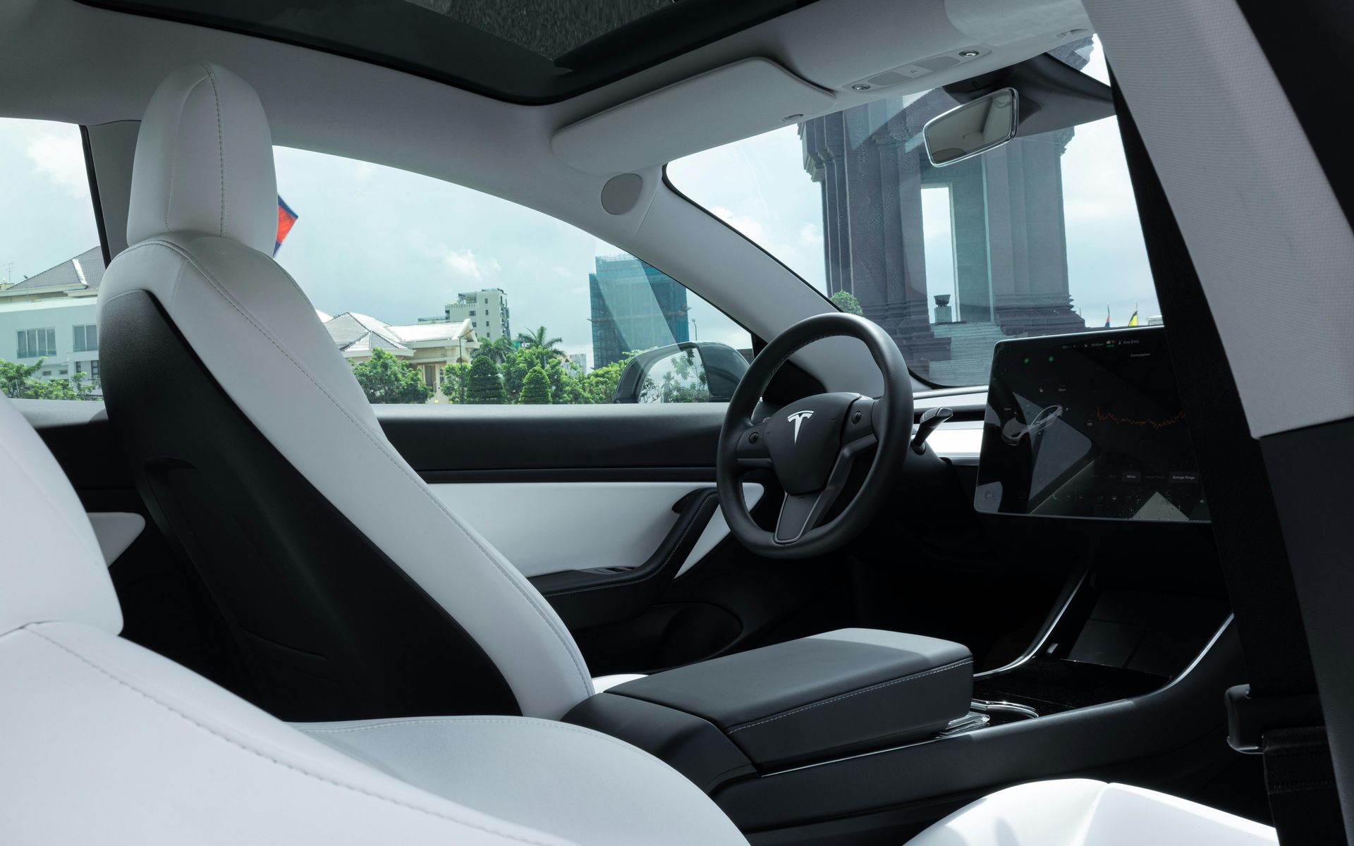 Interior of a Tesla car with white seats, black dashboard, and a large central touchscreen, city view outside.