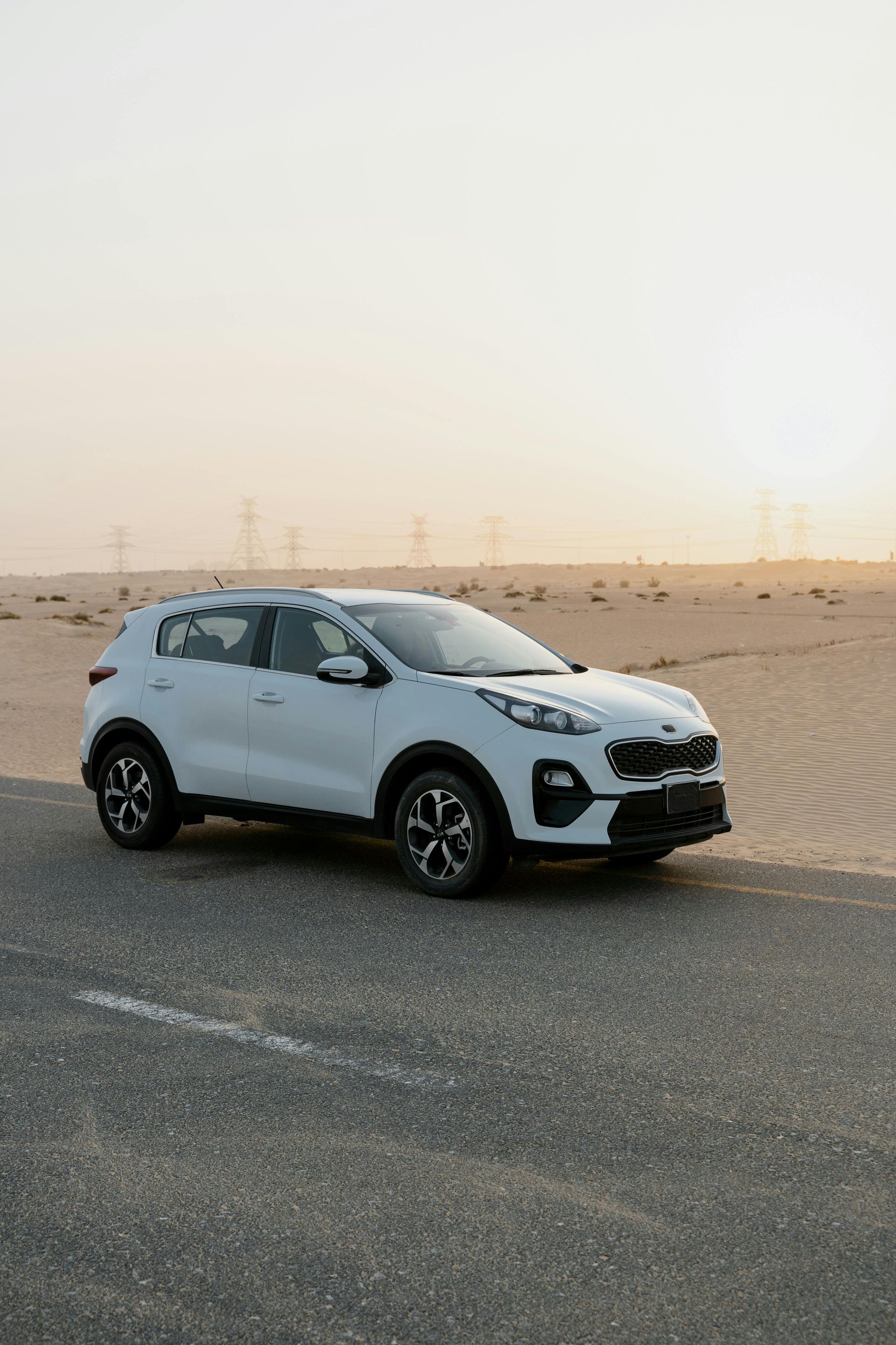 White Kia SUV parked on a road in a desert setting at sunset.