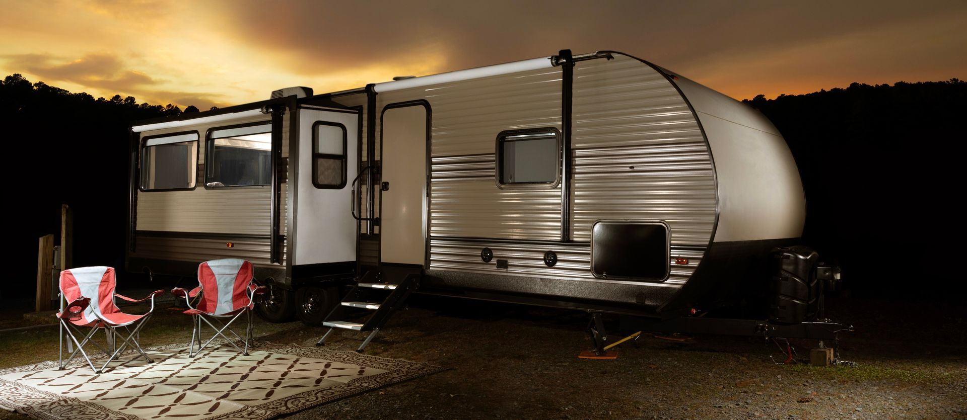 A camper at dusk with two red chairs lit by a light, set on a stone path.