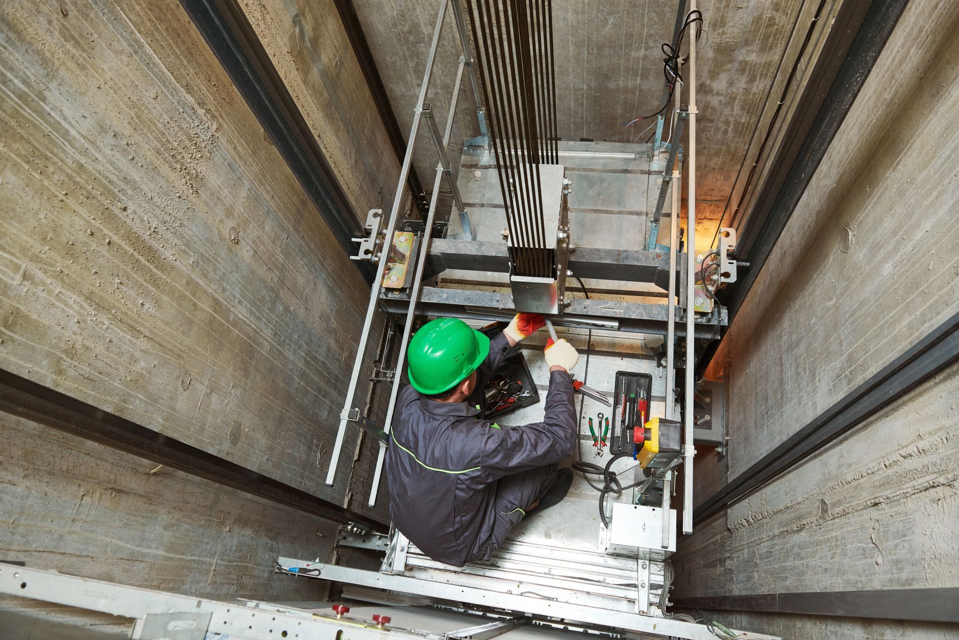 Un trabajador que viste un casco verde realiza tareas de mantenimiento en los cables y equipos del ascensor dentro de un hueco de ascensor de hormigón.