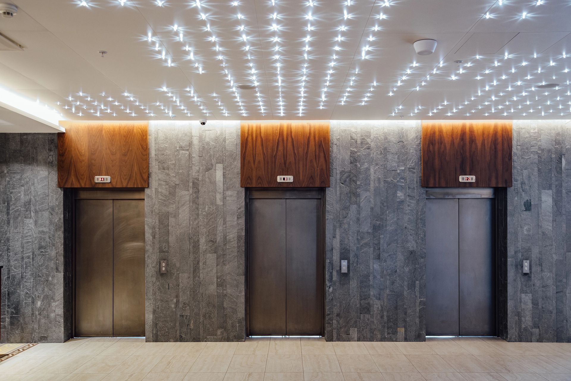 Un moderno vestíbulo de ascensor con tres puertas de acero inoxidable frente a paredes de piedra texturizada y un techo estrellado.