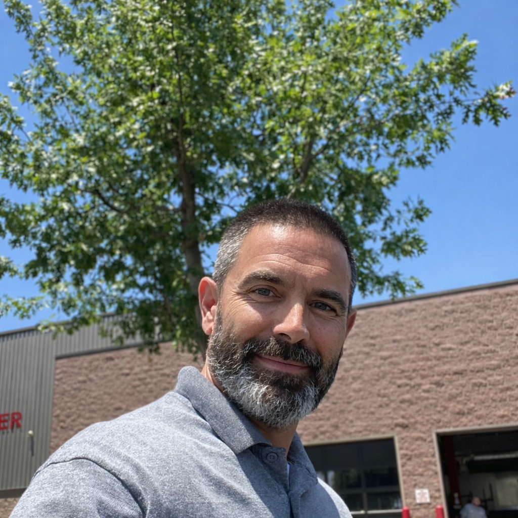 Man with a beard in front of a building and a tree, smiling, wearing a gray polo. Blue sky background.