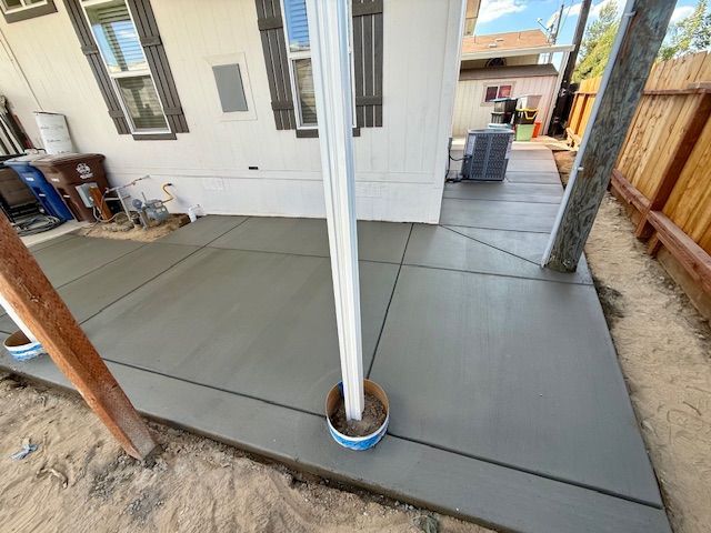 Newly poured gray concrete patio with control joints, next to a house and wooden fence.