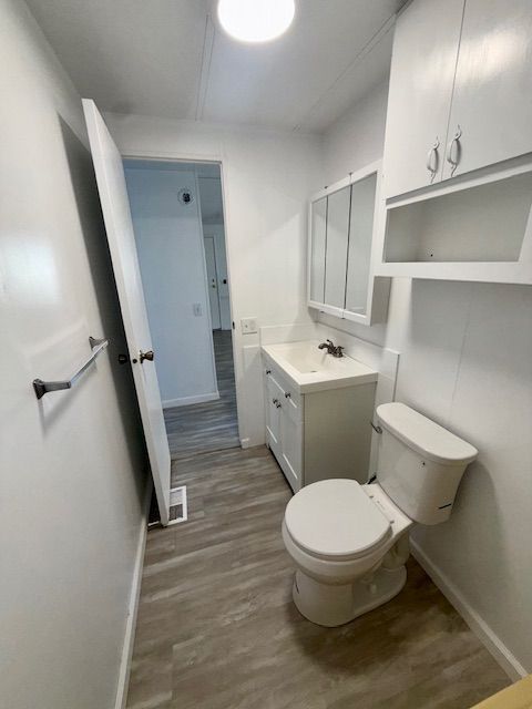 Small, white bathroom with toilet, sink, and cabinets. A partially opened door is leaning against the wall.