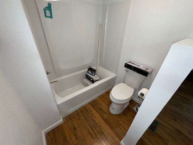 Small bathroom with a white tub and toilet, brown flooring, and white walls.