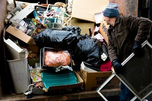 Person rummages in a dumpster filled with trash, holding a window screen.