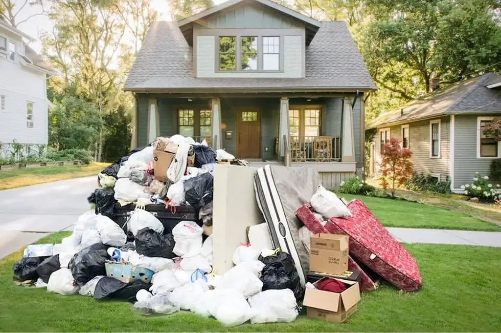 Pile of trash and discarded items on front lawn of a house; next to a sidewalk and other houses.