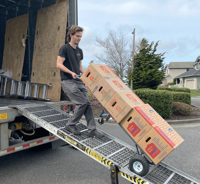 Person loading cardboard boxes onto a truck using a ramp and hand truck. Outdoors, day.