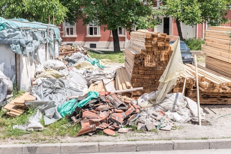 Construction debris: piles of wood planks, rubble, and covered waste, in front of a red brick building.