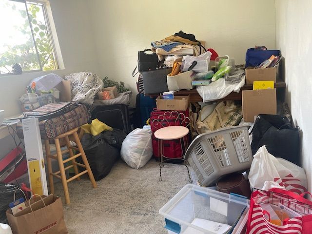 Room filled with disorganized piles of boxes, bags, and items; light shines in from a window.