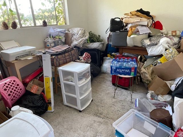 Room cluttered with various items: boxes, clothing, plastic bins, a printer, and other objects.