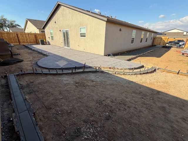 Backyard with unfinished patio construction; gray pavers, concrete border, dirt ground, house in the background.
