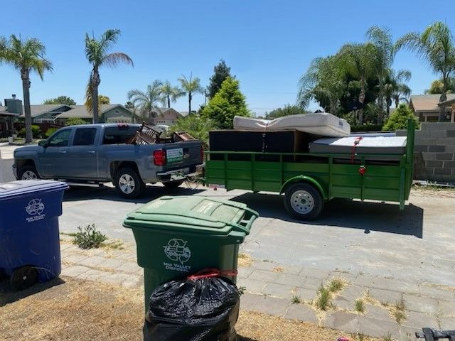Gray pickup truck towing a green trailer filled with furniture, parked near trash bins in a residential area.