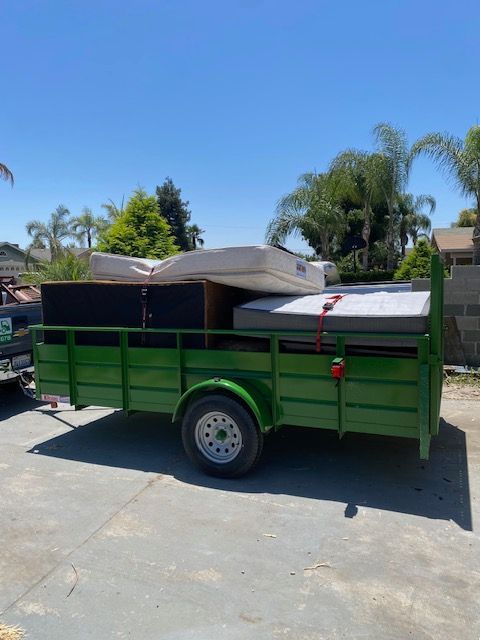 Green trailer loaded with mattresses and a large black box, parked outdoors on concrete, sunny day.