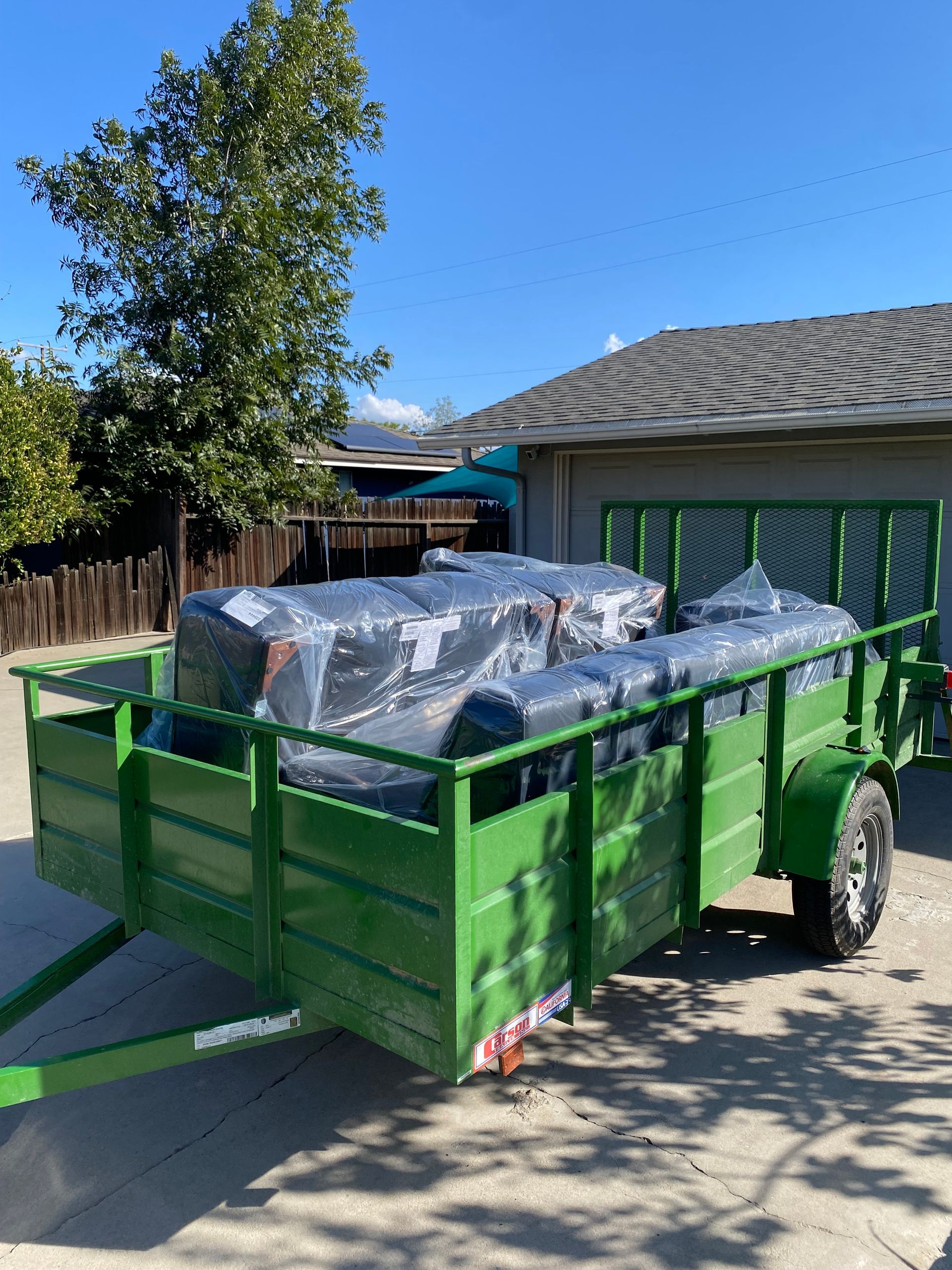 Green trailer loaded with covered items parked in front of a house on a sunny day.