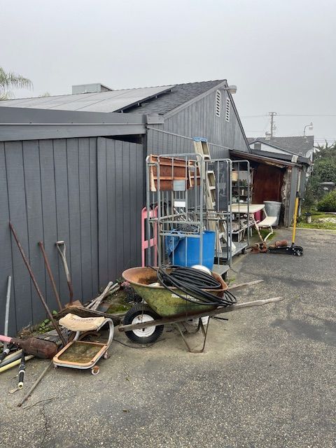 Assorted tools and objects cluttering a driveway next to a gray wooden shed, a red shed, and a roof with solar panels.