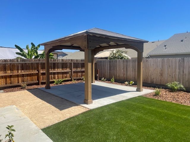 Wooden gazebo on concrete patio, grass lawn, gravel, wooden fence, sunny day.