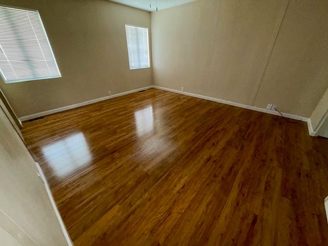 Empty room with hardwood floors, two windows with blinds, and tan walls.
