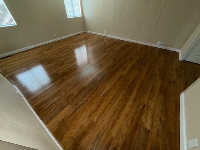 Hardwood floor in an empty room, reflecting light from windows. Beige walls, white trim.