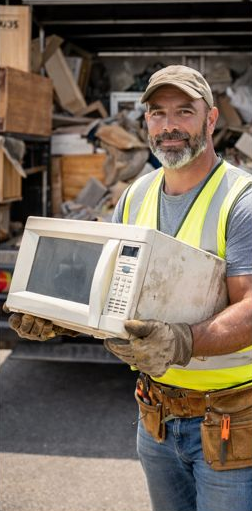 Man in work clothes holding a microwave in front of a truck filled with trash.