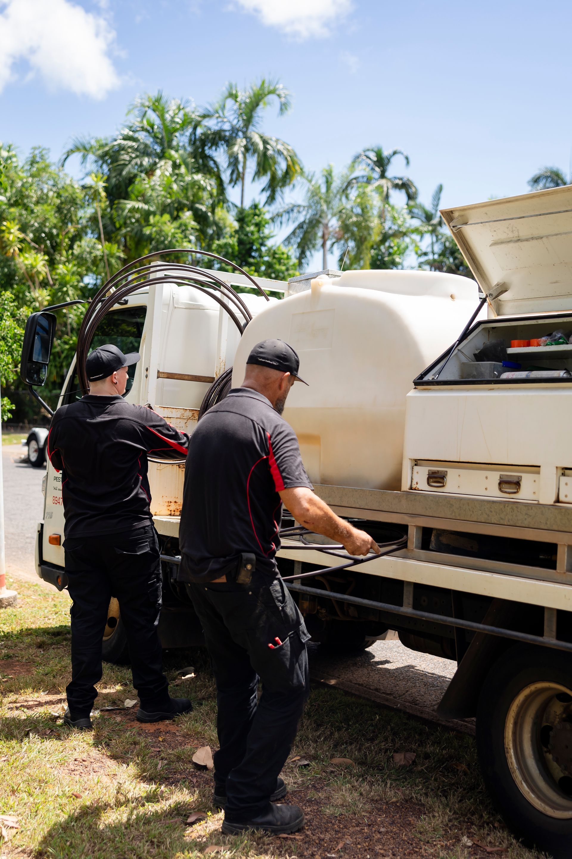 Two Men in Black Shirts and Hats Are Working on a White Truck Outdoors — Quack Pest Control in Parap, NT