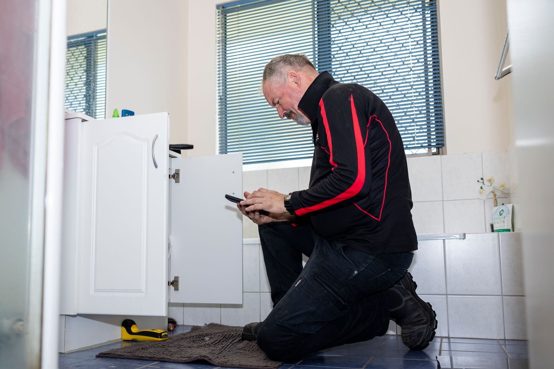 Man Kneeling in Bathroom, Looking at Phone While Working on a Cabinet, Near Window — Quack Pest Control in Nightcliff, NT