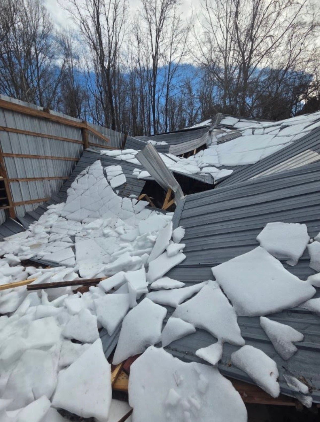A collapsed metal roof on a structure with broken wooden beams, covered in large chunks of ice and snow.