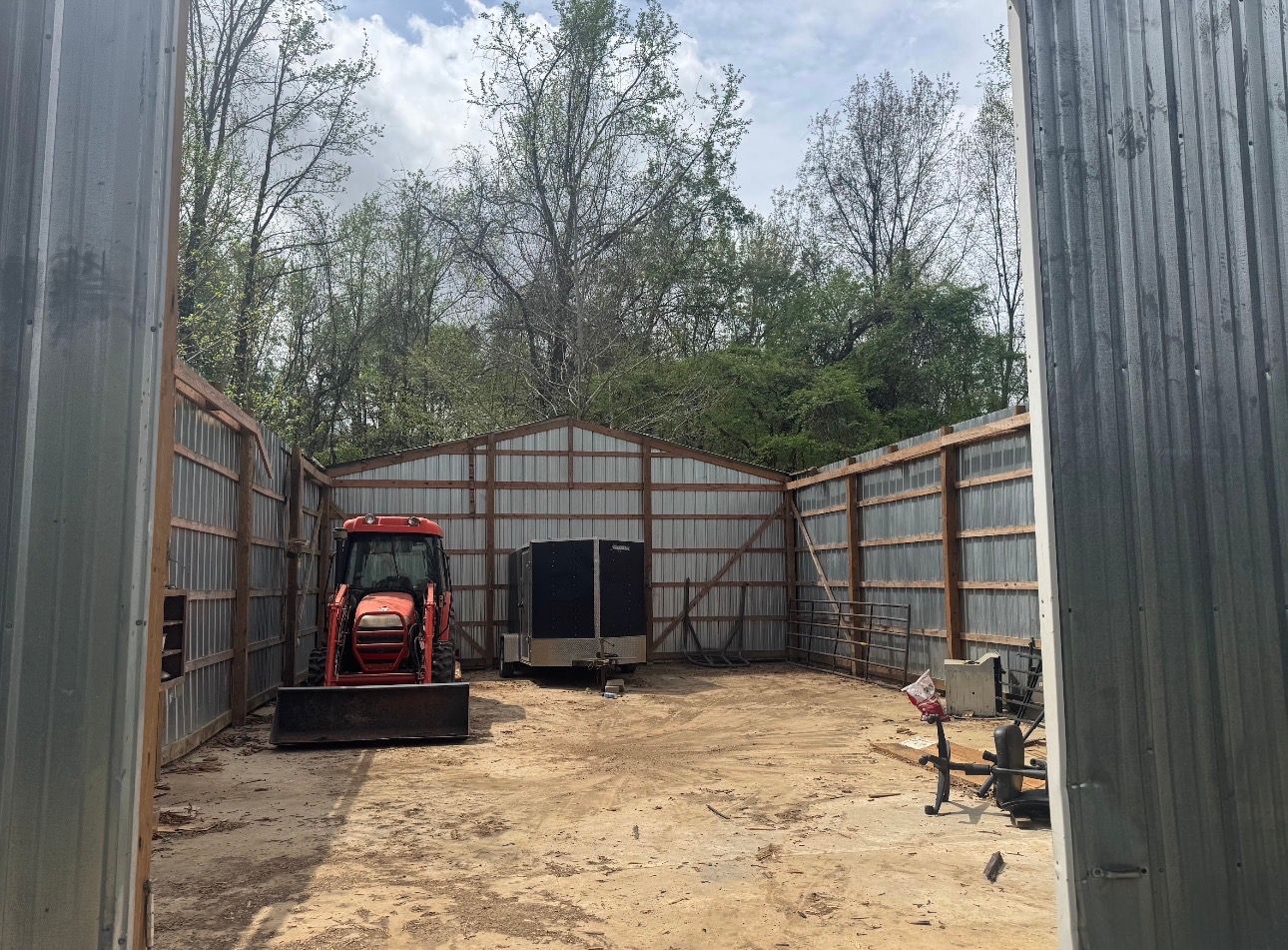 An open-ended metal barn containing a tractor and a small black trailer on a dirt floor, surrounded by trees.