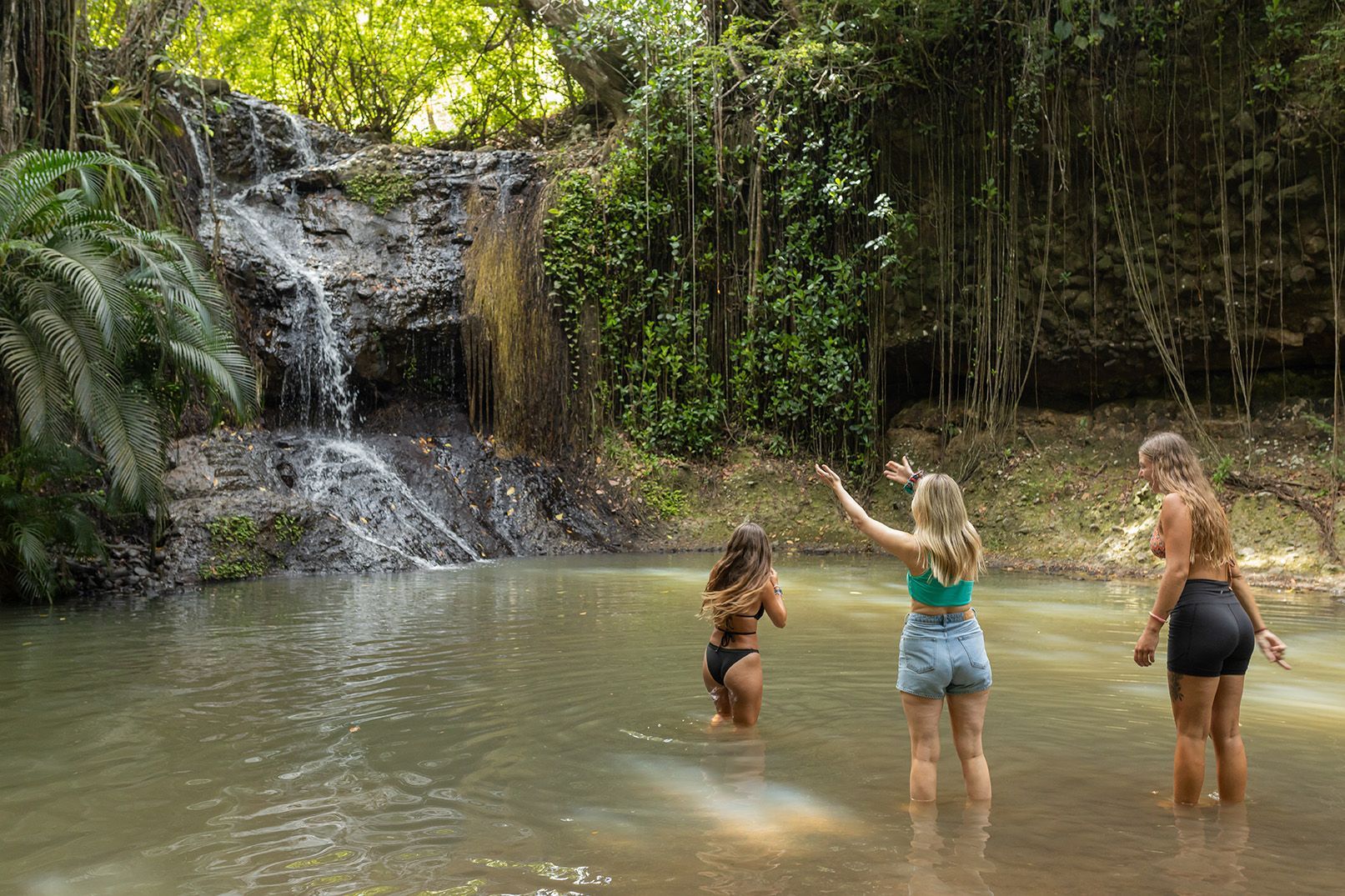 Three women are standing in a pond near a waterfall.