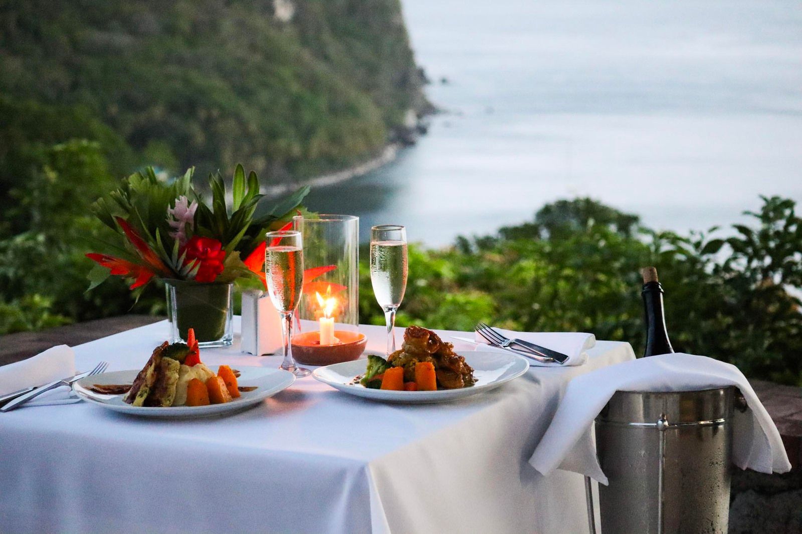 A table with two plates of food and two glasses of wine with a view of the ocean.
