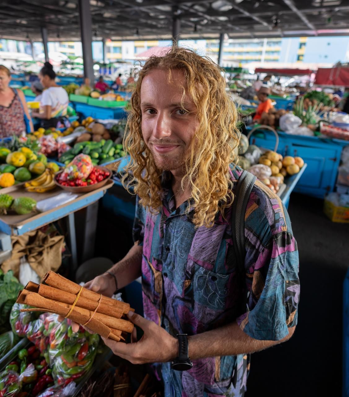A man with long curly hair is holding a bunch of carrots in a market.