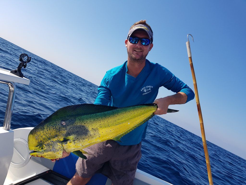 Man on boat holds up large, colorful Mahi-Mahi fish he caught in the ocean; blue sky.
