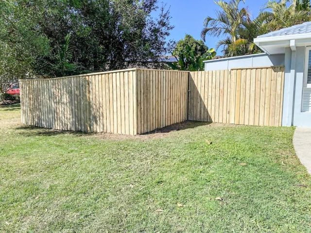 Wooden fence encloses a green lawn, near a house with a gray exterior on a sunny day.