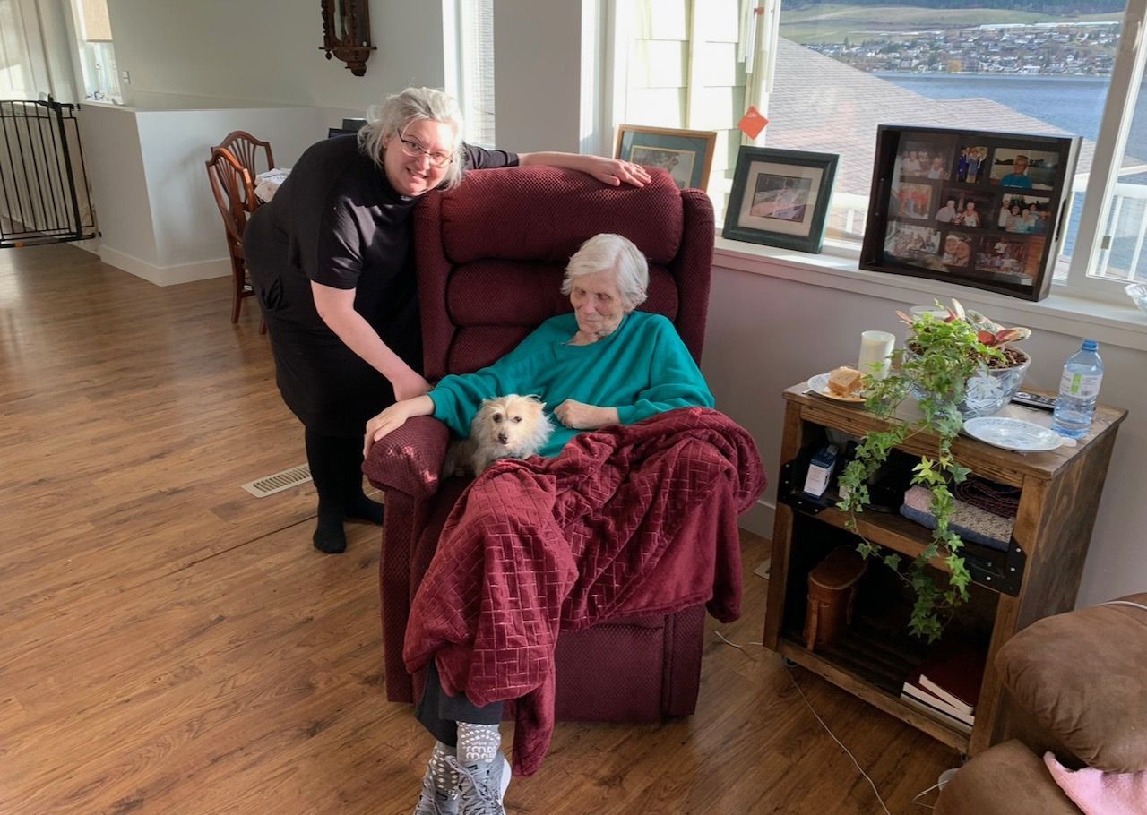 A woman is standing next to an elderly woman sitting in a chair with a dog.