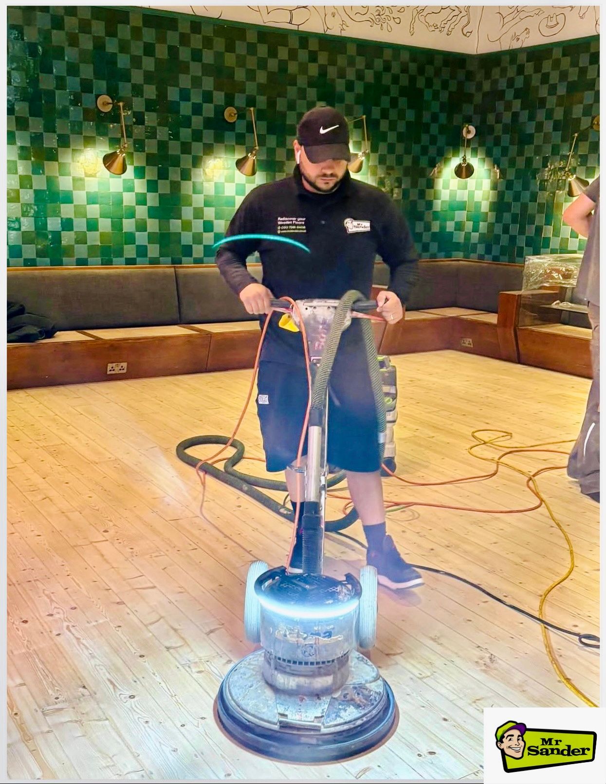 Man sanding wooden floor with a floor buffer in a room with green tiled walls.