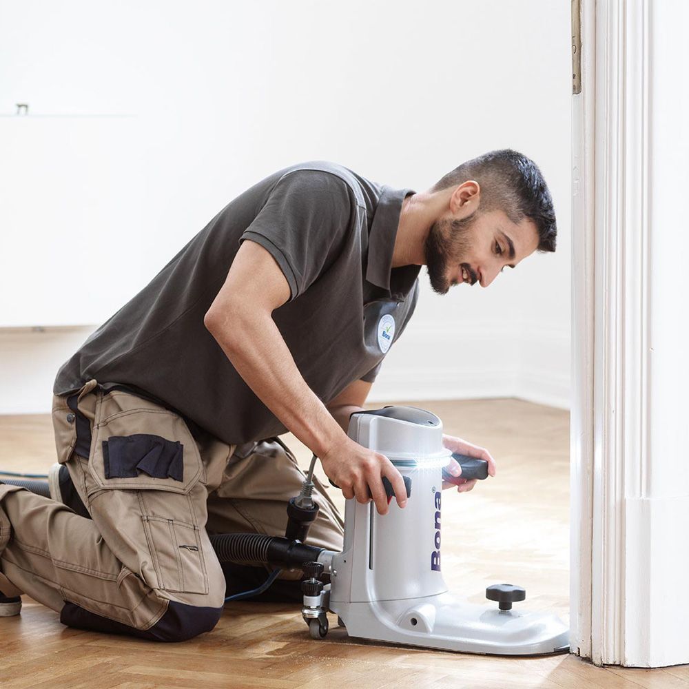 Man kneels, operating a floor sander next to a door. He wears a gray shirt and tan pants; the setting is indoors.