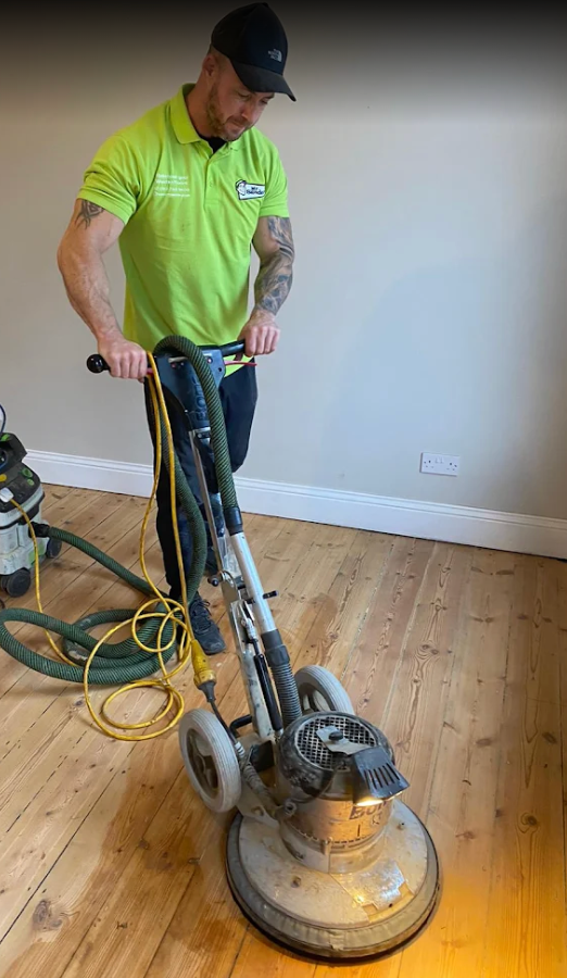 Person in green shirt using a floor sander on wooden planks, interior setting.