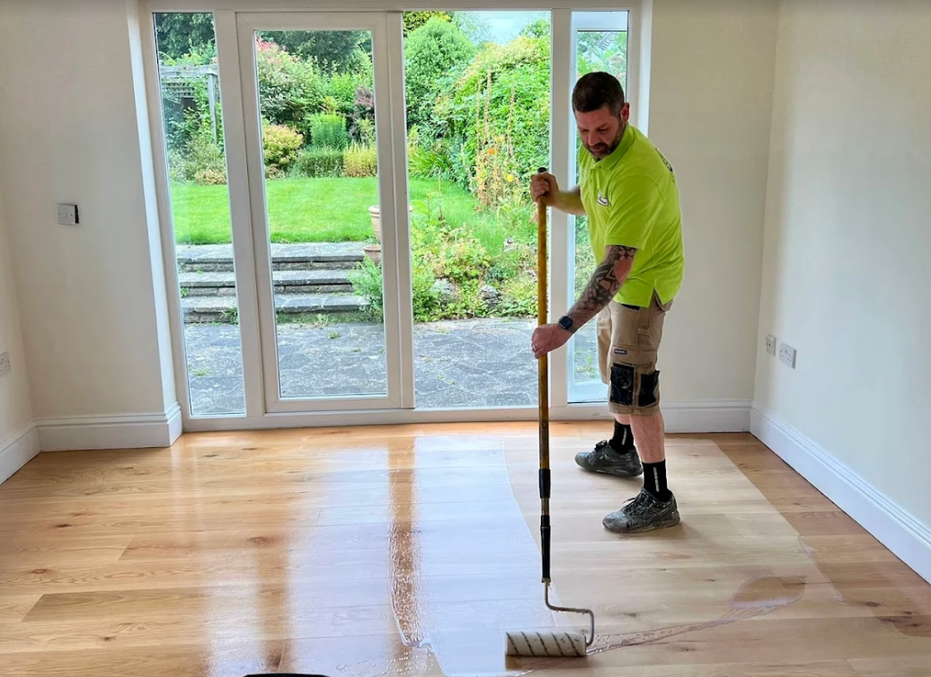 Man applying sealant to a wooden floor with a roller. Interior room with large windows, bright wood, and beige walls.