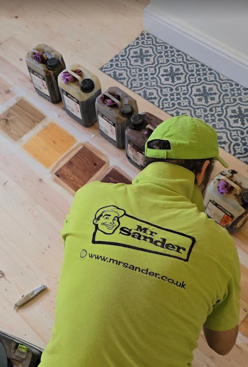 A person in a green shirt with the "Mr. Sander" logo is applying stain to wood flooring.