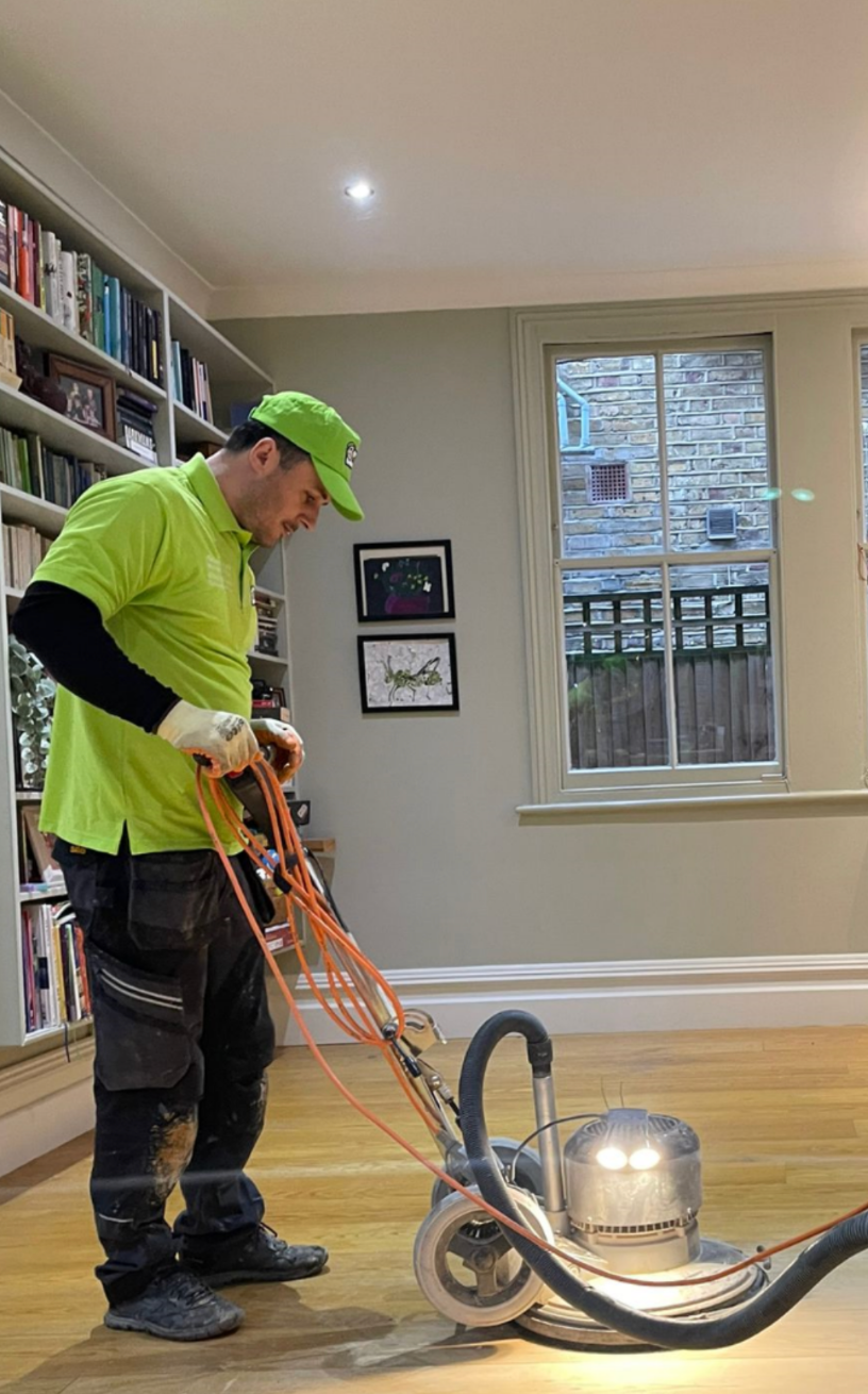 Man in green shirt using a floor sander on hardwood floor in a room with a bookcase and window.