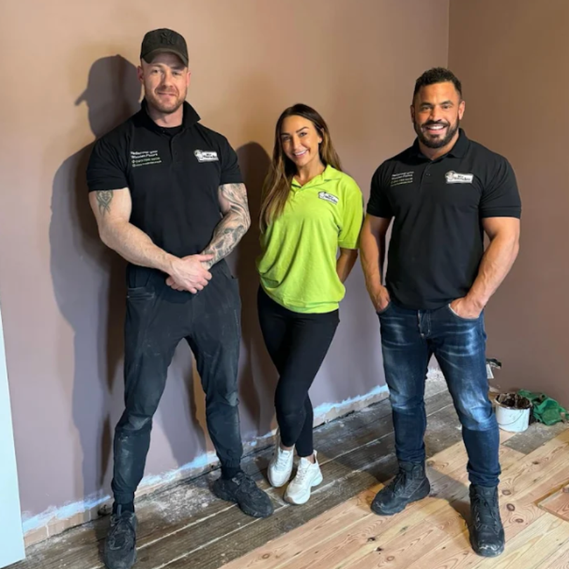 Three people posing in a room with exposed flooring. Two men in black shirts and a woman in a green shirt.