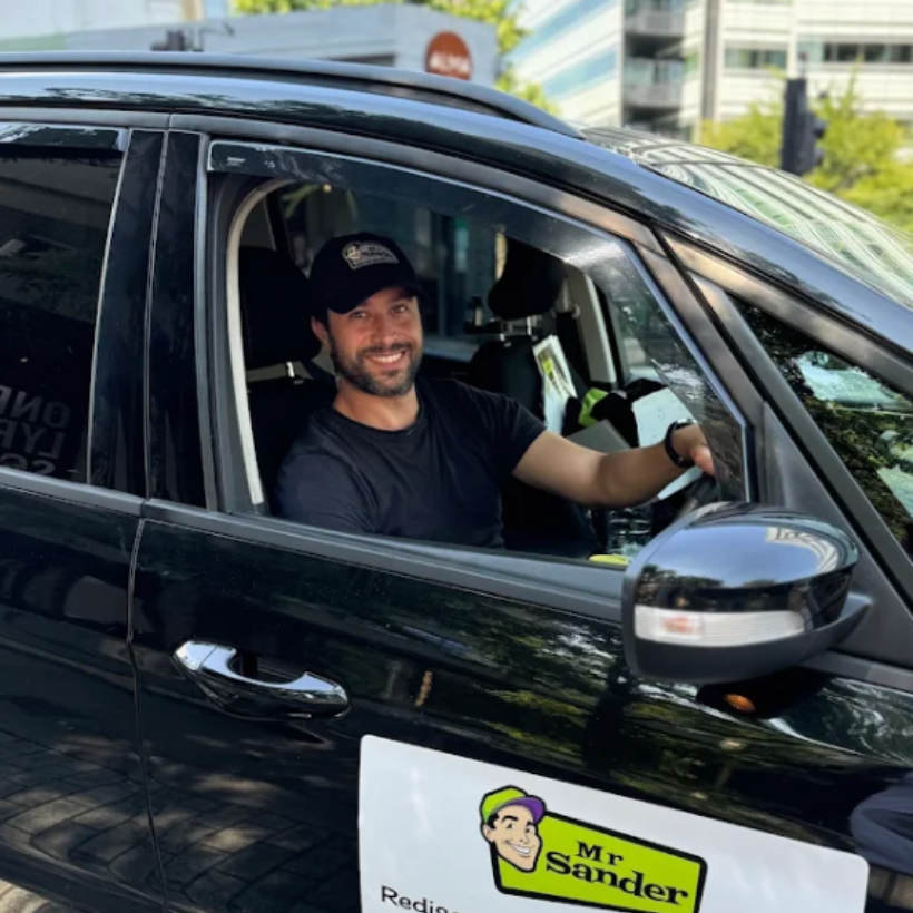 Man in black vehicle, smiling, wearing a hat with a logo. A business sign is visible on the vehicle.