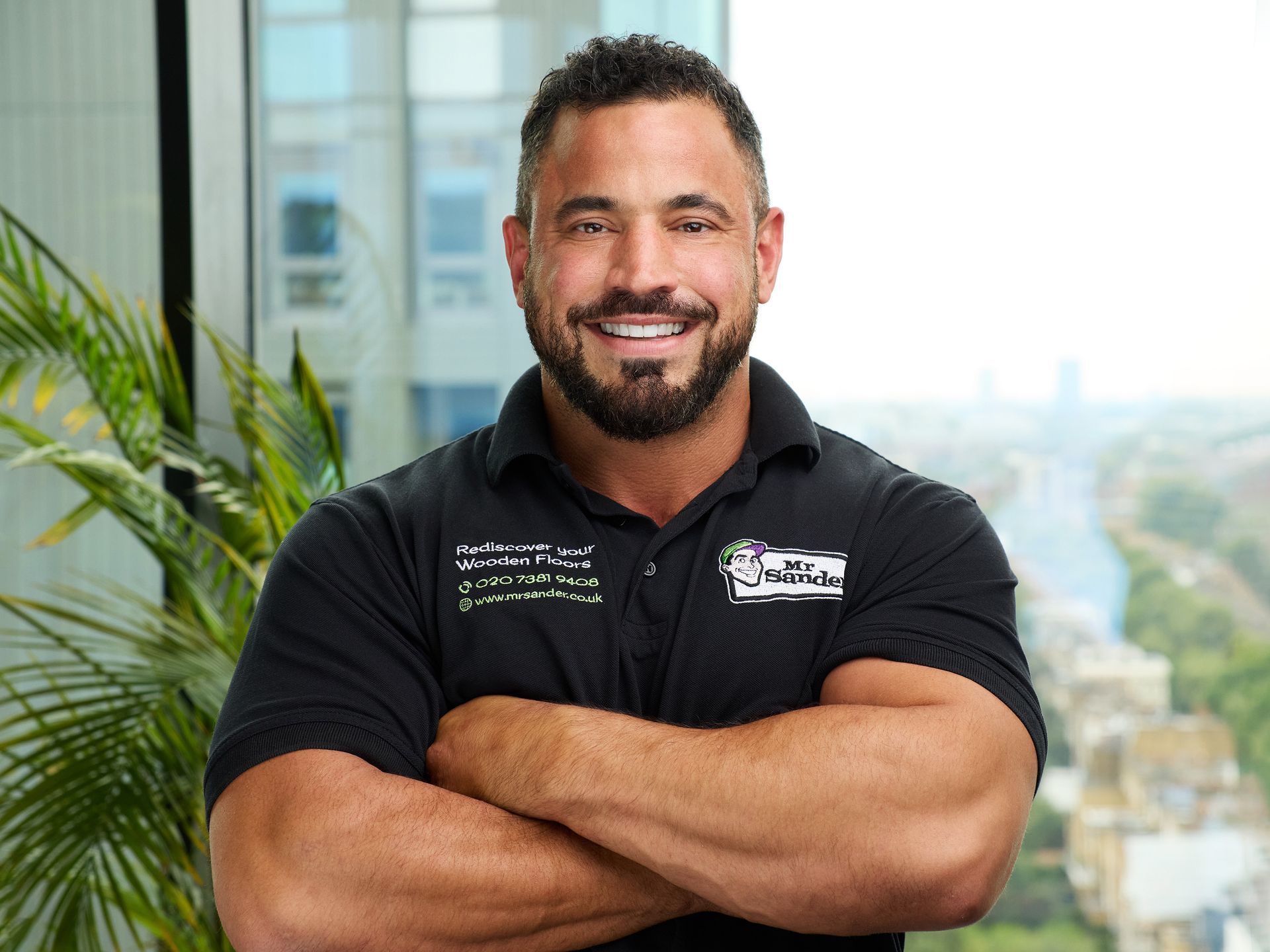 Man with a beard and crossed arms, wearing a black shirt, smiles. City background.