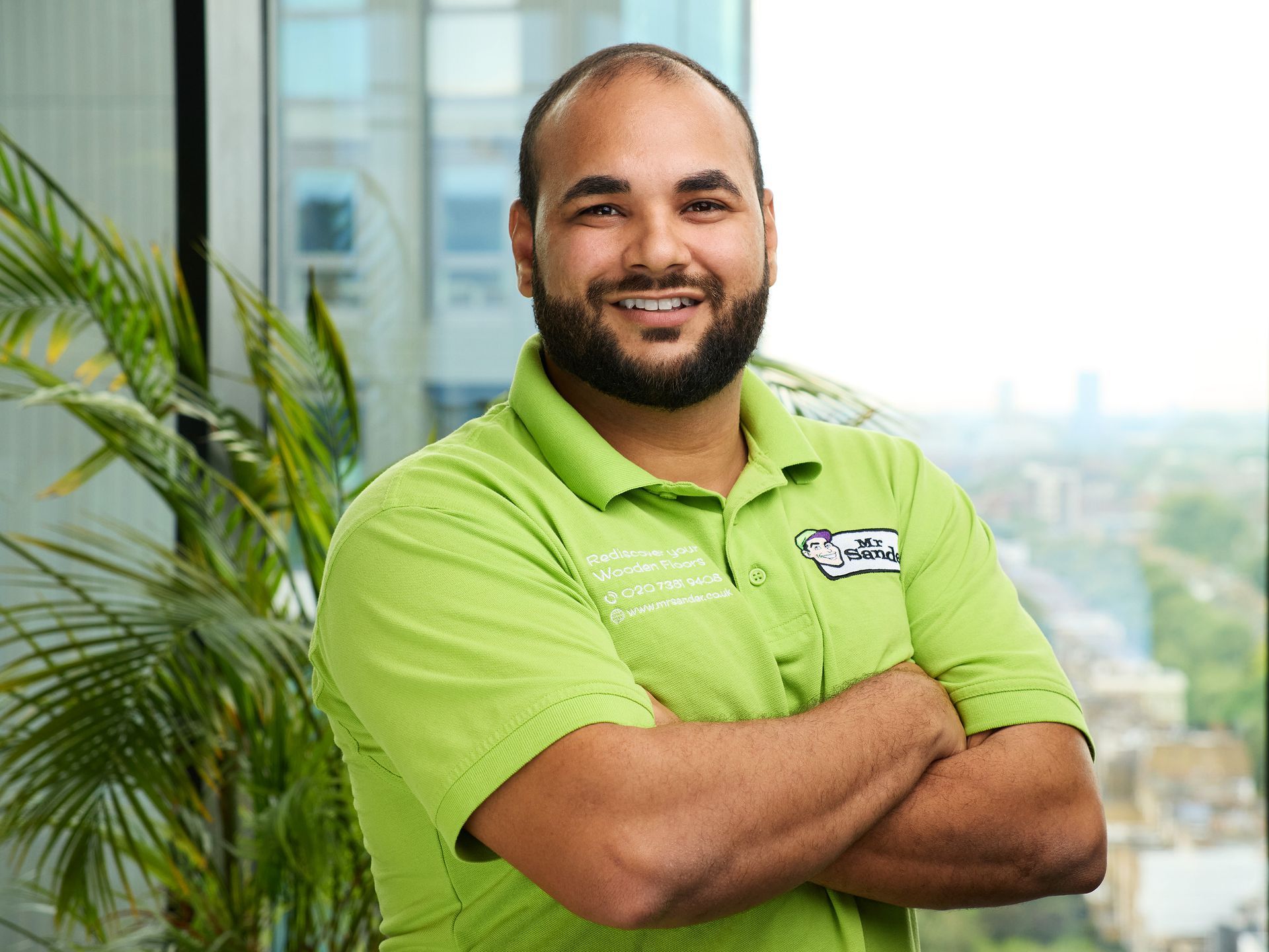 Man in green shirt, arms crossed, smiling. Blurred cityscape in background, indoor setting.
